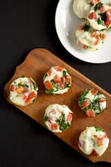 Homemade Egg White Breakfast Cups with Spinach and Tomato on a rustic wooden board on a black background, top view. Flat lay, overhead, from above.
