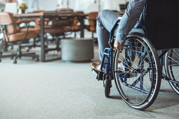 A disabled man is sitting in a wheelchair. He holds his hands on the wheel. Nearby are his colleagues. Disabled businessman wearing suit sitting in a wheelchair in an office