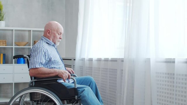 Disabled old man is sitting in a wheelchair at home alone. A handicapped person in a wheelchair is looking out the window. Paralysis and disability.