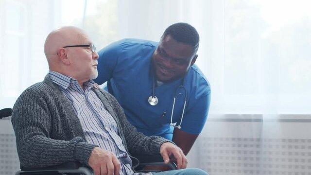African-American caregiver and old disabled man in a wheelchair. Professional nurse and handicapped patient in a nursing home. Assistance, rehabilitation and health care.