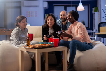 Multi ethnic diverse group of friends playing fun game on tv console after work at office. Cheerful colleagues holding joystick controller for gaming indoors on sofa. Happy team celebrating