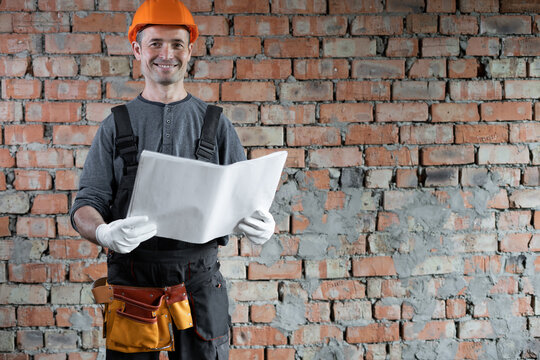 smiling builder with a plan or drawing in his hands standing near a brick wall in the house. An adult engineer in a protective construction helmet. Repair and construction concept.