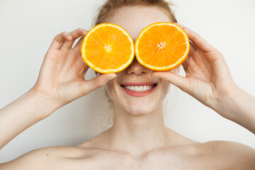 smiling ginger woman with freckles is covering her face with sliced oranges posing on a white wall