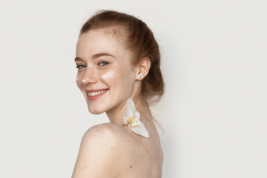 Back view photo of a ginger woman with freckles smiling at camera on a white wall applying anti aging cream on face and holding a flower on shoulder
