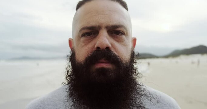 Serious latin man with big beard on a beach looking at horizon. 4K.