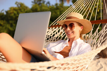 Fototapeta premium Working at sunset. Side view of young happy female using laptop while lying in the hammock on the beach