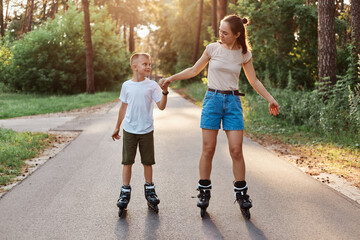 Fototapeta premium Outdoor full length portrait of happy brunette female holding son's hand and rollerblading together, family wearing casual style clothing, spending time in park.