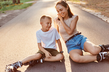 Fototapeta na wymiar Mother and son in roller skates sitting on a road, mommy talking with her little boy, enjoying beautiful nature and fresh air, active weekends together.