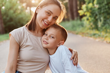 Fototapeta premium Portrait of mother and son smiling and hugging each other and looking at camera, happy family outdoor, having fun together in summer park, childhood, motherhood.
