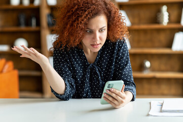 Shocked red-haired curly woman holding a smartphone, feel astonishment with a bad news, fired from...