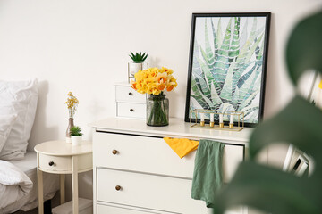 Chest of drawers with narcissus flowers near white wall in bedroom