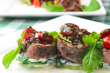 Grilled meat steaks with fresh vegetables and sauce served on a white square plate over green plaid tablecloth.