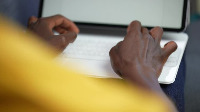 Closeup POV shot of black afro-american man's hands typing on a white laptop or tablet keyboard resting on his knees. 