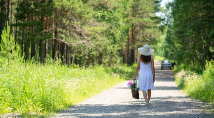 A beautiful girl in a white sundress and a hat is walking along a forest road with peonies in her hands. The concept of a summer holiday.