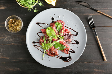 Plate with tasty veal carpaccio and ingredients on dark wooden background