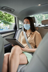 Woman wearing protective mask is sitting inside the car and using tablet with interest