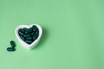 Green capsules for skin in heart shape bowl, on green background. Woman's care, detox program....
