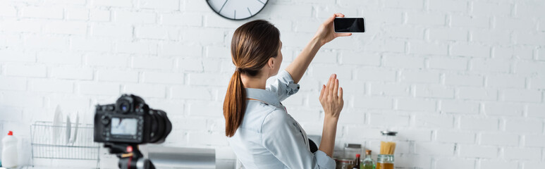 housewife waving hand near smartphone with blank screen and blurred digital camera in kitchen, banner.