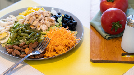 Healthy mixed colorful salad with mixed greens, carrot, almonds, tomatoes, eggs, black olives and meat. Wood serving board with green napkins and two tomatoes. Yellow background. Fresh vegetable salad