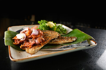 grilled grouper fish fillet with spicy onion sambal chilli sauce on banana leaf in dark background asian halal seafood menu