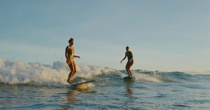 Beautiful girls surfing at sunset, best friends riding wave together, active lifestyle outdoors, epic slow motion
