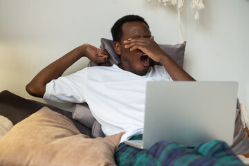 African man working lying on bed at home being tired yawning.