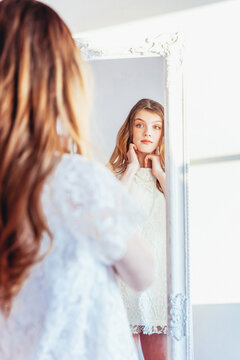 Beauty Makeup Morning Rutine Love Yourself Concept. Young Teenage Girl Looking At Reflection In Mirror. Young Positive Woman Wearing White Dress Posing In Bright Light Room Against White Wall.