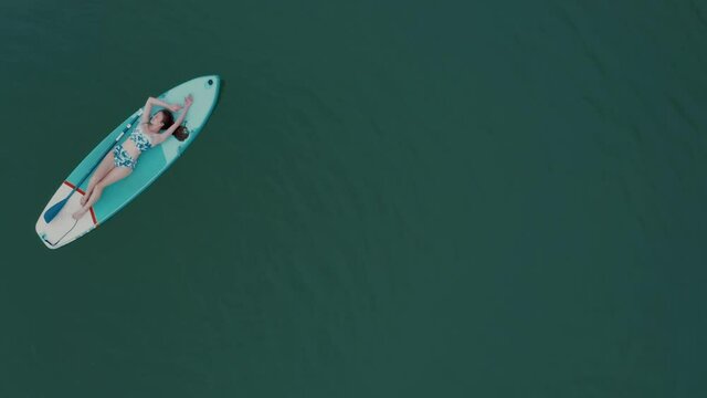 Young woman in bikini lying on sup board with paddle on water surface top view. Surfer girl relaxing, tanning on inflatable board for rowing, float, chill in ocean, summer vacation, surfing activity