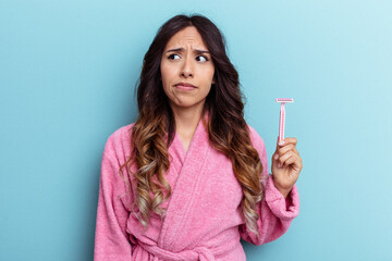 Young mexican woman wearing a bathrobe holding a knife isolated on blue background confused, feels doubtful and unsure.