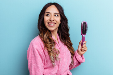 Young mexican woman wearing a bathrobe holding a brush isolated on blue background looks aside smiling, cheerful and pleasant.