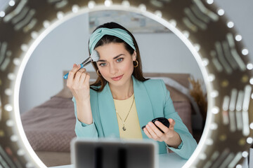 blurred smartphone in holder with circle light near beauty blogger applying face powder.