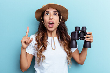 Young mexican woman holding binoculars isolated on blue background pointing upside with opened mouth.