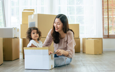 Happy beautiful Asian mother with little Caucasian cute daughter laughing, playing together to put the girl in paper box and push with fun and happiness while moving to new house. Relocation concept.