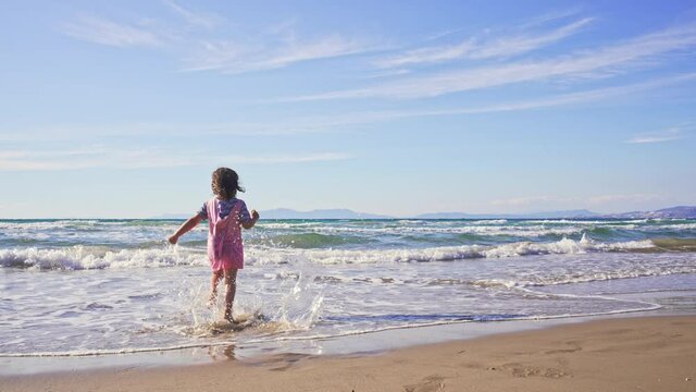 Little girl run into the sea water, beautiful sunny day. Happy child playing on the beach with waves.