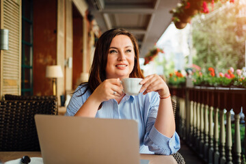happy woman sits with laptop in cafe on street and drinks coffee