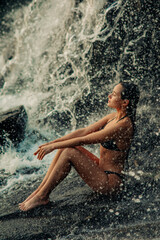 Fototapeta premium Beautiful woman sits under the waterfall and enjoys the falling water.