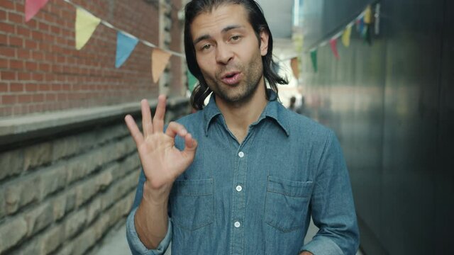 Slow motion of optimistic mixed race man making ok hand gesture smiling and saying okay outdoors in city street. Modern people and attitude concept.