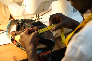 African tailor measuring with a measure tape, near by a sewing machine at sewing workshop. Black...
