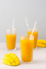 Freshly squeezed mango juice in glasses with a straw, ripe cut mango on a gray background