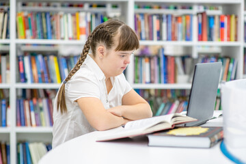 Pensive Young girl with syndrome down uses a laptop at library. Education for disabled children concept