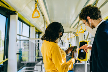 Rear view of multi ethnic mixed race couple in protective masks buy ticket with smartphone and ticket machine in modern bus during ride. Communication, acquaintance, friendship concept.