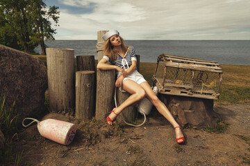 Young blonde woman with crimped hair, dressed in a sailor style outfit, sitting on wooden pillars...