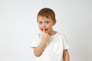 Cute little boy in casual clothing standing on white background of studio and putting index finger on lips while looking at camera. The child keeps a secret