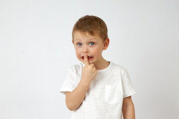 Fototapeta premium Cute little boy in casual clothing standing on white background of studio and putting index finger on lips while looking at camera. The child keeps a secret