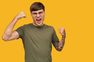 Indoor shot of young male, wears green t-shirt and round stylish spectacles, shouting with widely...