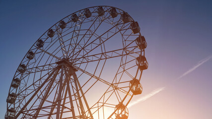 Large operating metal Ferris wheel amusement ride rotates in local park under clear blue sky in spring evening