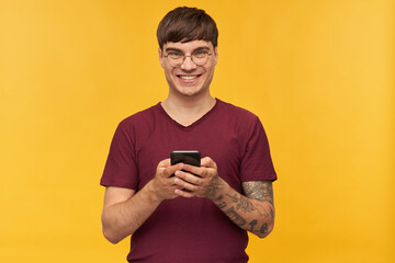 Indoor shot of young happy smiling male, looks directly into camera while typing a post in social network, wears red t-shirt and round stylish glasses. Isolated over yellow background