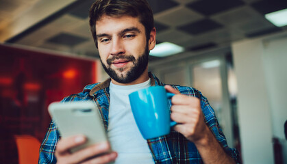 Young Caucasian male blogger holding coffee mug enjoying break for online messaging and chatting, millennial hipster guy with tea cup reading received email browsed on smartphone technology - obrazy, fototapety, plakaty