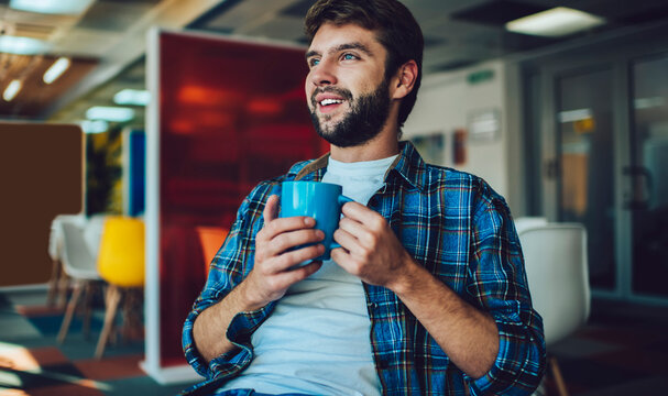 Young Caucasian male coworker dressed in shirt holding coffee mug enjoying break for thinking and dreaming indoors, millennial hipster guy with tea cup feeling pondering during day in workshop space