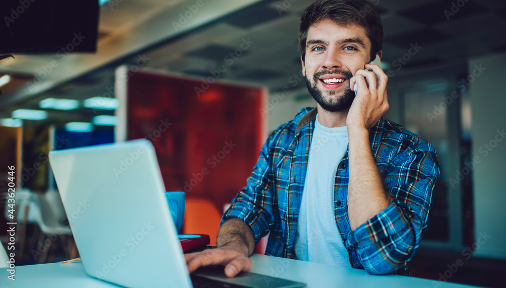 Poster portrait of happy caucasian hipster guy smiling at camera while using cellular for positive calling 